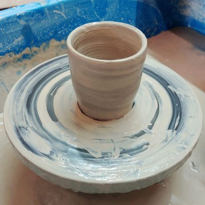 beige cup shaped clay cylinder on pottery wheel head with fresh slip marks covering most of the wheel and splash tray showing blue plastic frame in the top half of the image